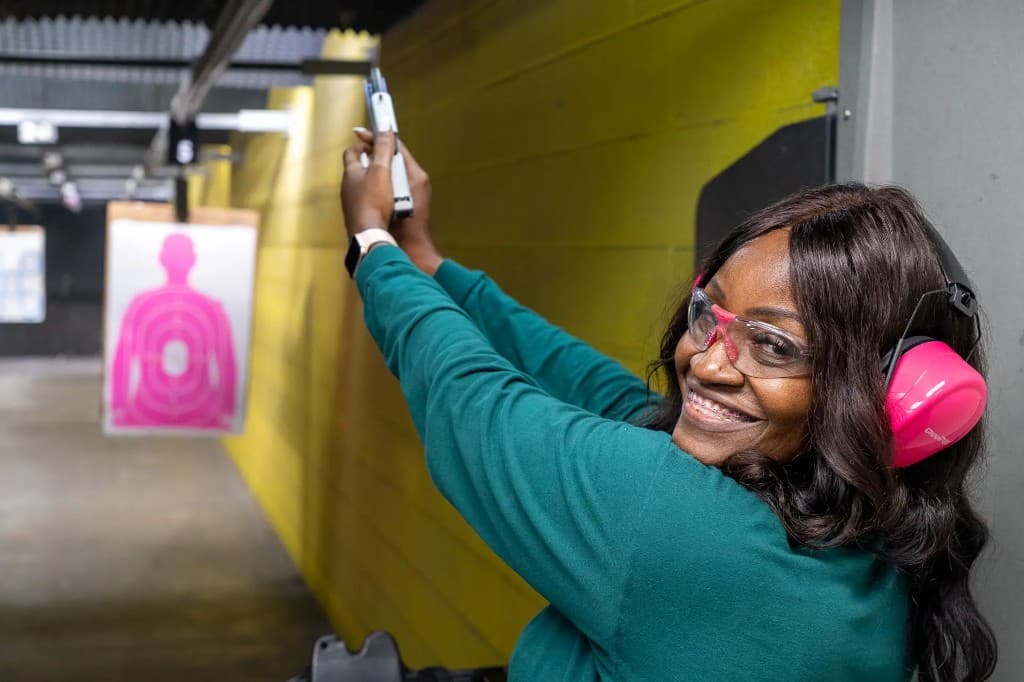 Shooter on an indoor range lane with eye and ear protection, aiming downrange toward a paper target